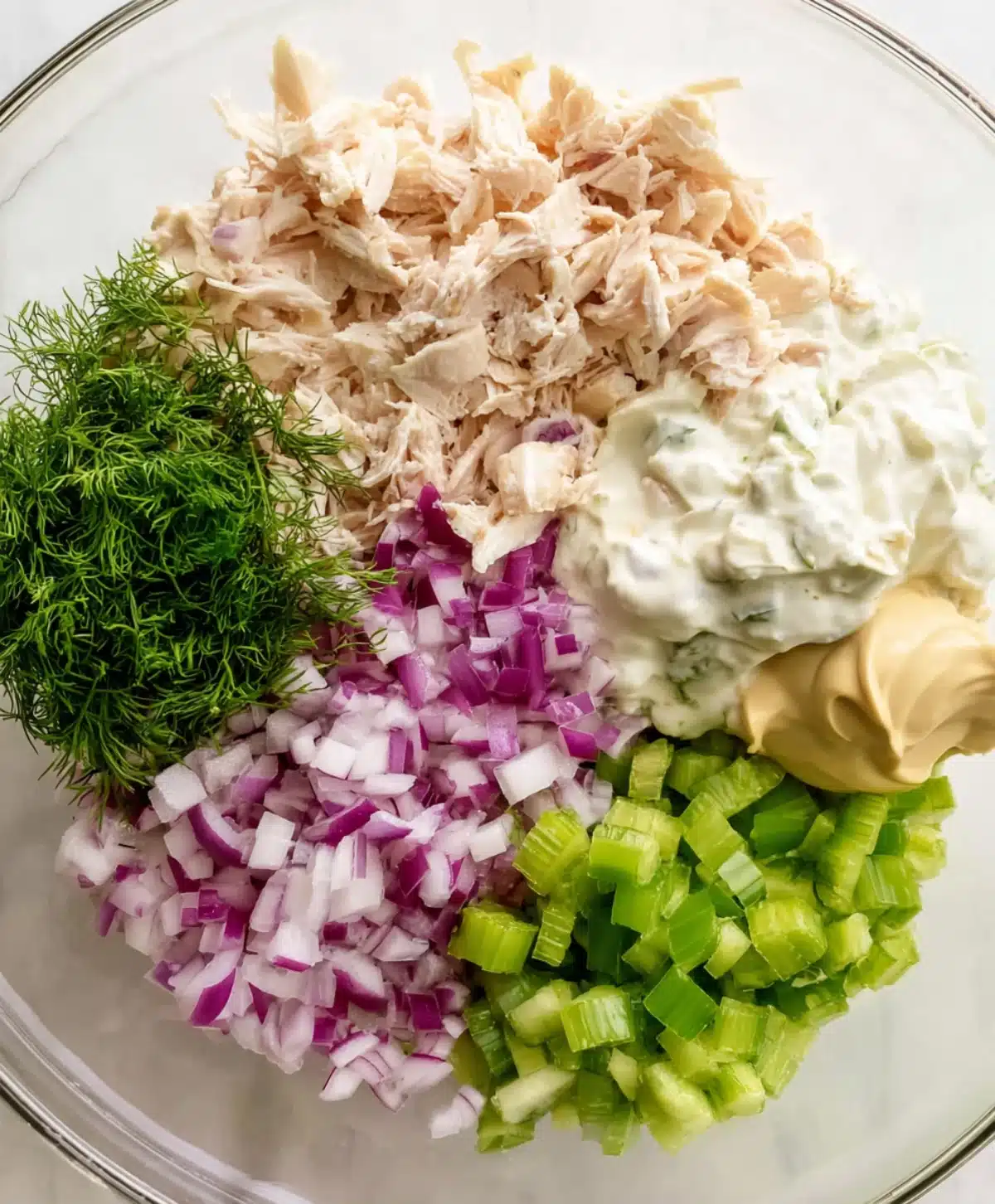 Ingredients for canned chicken salad including celery, red onion, dill, mayo, mustard, and shredded chicken in a bowl.
