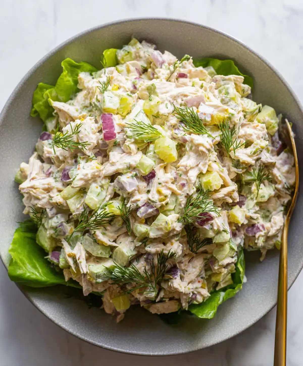 Canned chicken salad served on fresh lettuce leaves with red onion, pickles, celery, and dill in a grey bowl.