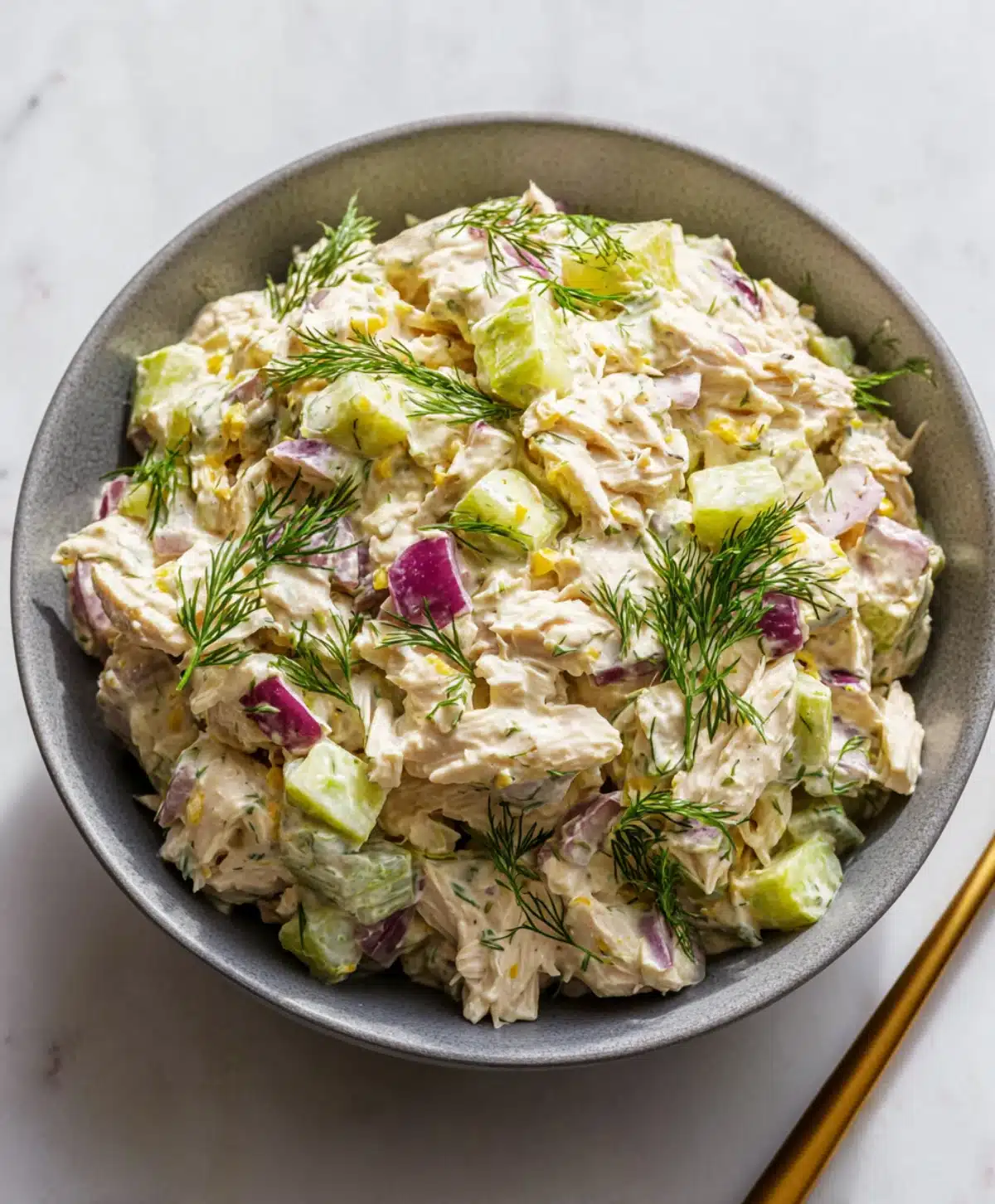 Bowl of creamy canned chicken salad topped with fresh dill, red onion, celery, and pickles on a marble surface.