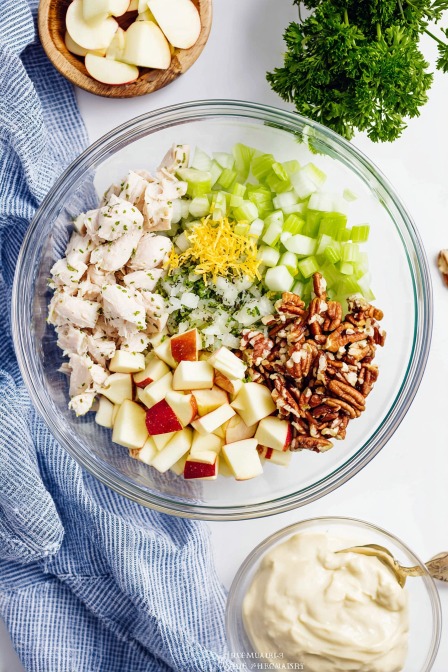Ingredients for chicken salad with apples, pecans, celery, lemon zest, and mayo arranged in a glass bowl ready to mix.