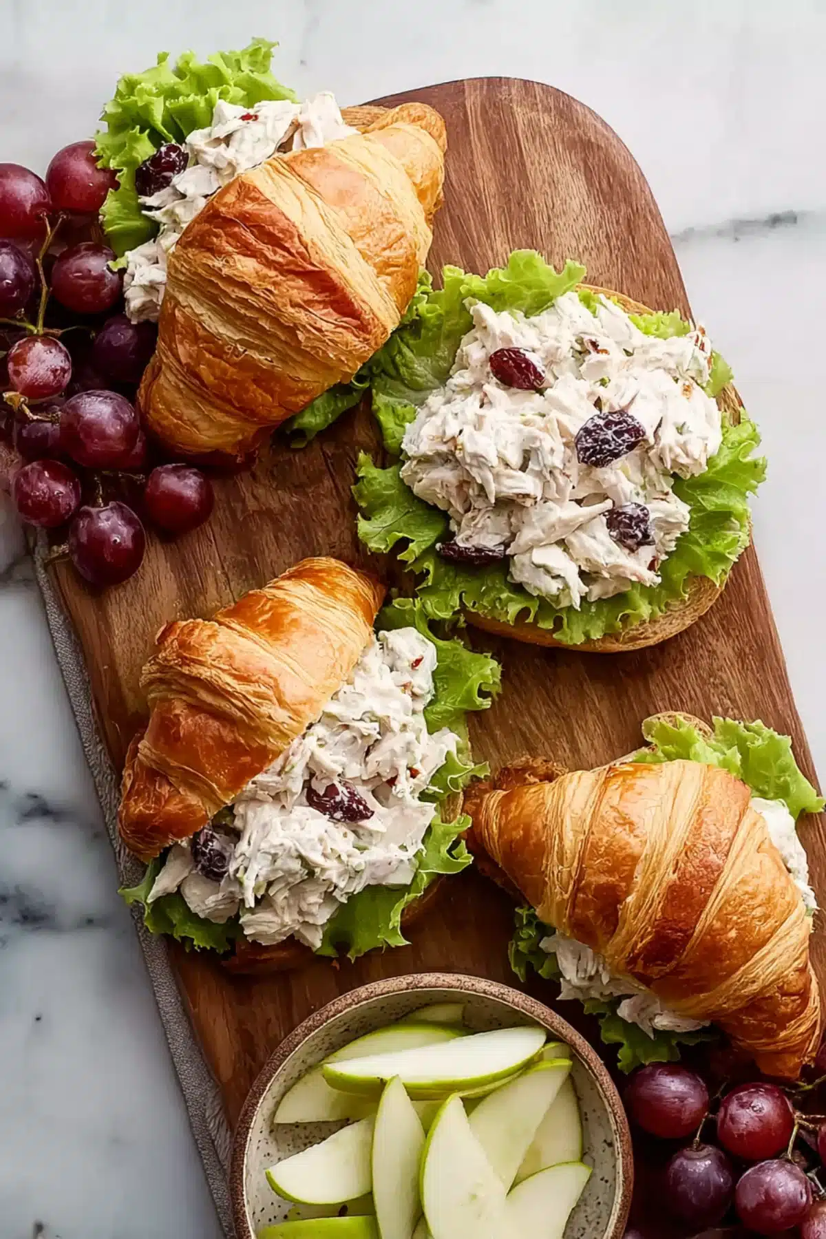 Chicken salad with grapes and lettuce served in croissants on a wooden board with sliced apples and fresh grapes.
