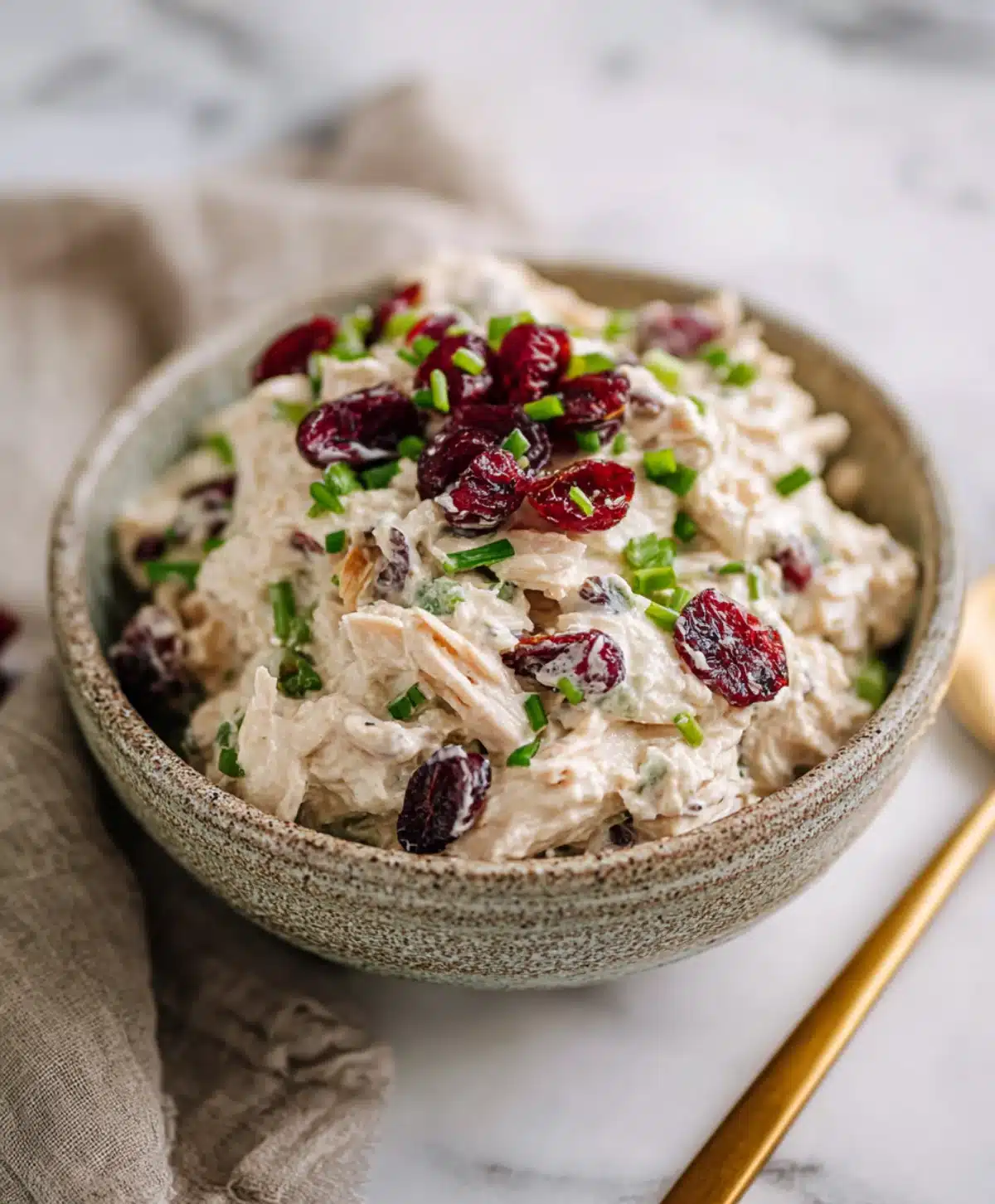 Creamy chicken salad topped with dried cranberries and chopped chives in a rustic bowl on a marble counter