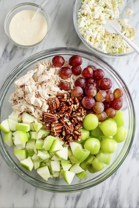 Ingredients for chicken salad including shredded chicken, apples, grapes, pecans, and celery in a glass mixing bowl
