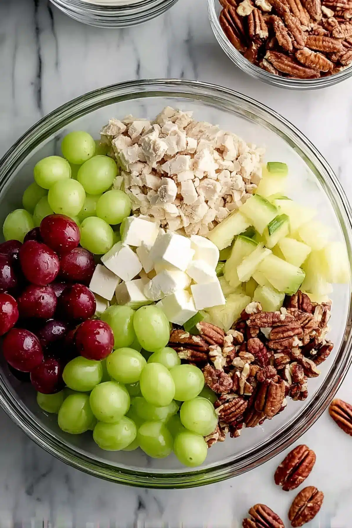 Ingredients for chicken salad including shredded chicken, apples, grapes, pecans, and celery in a glass mixing bowl