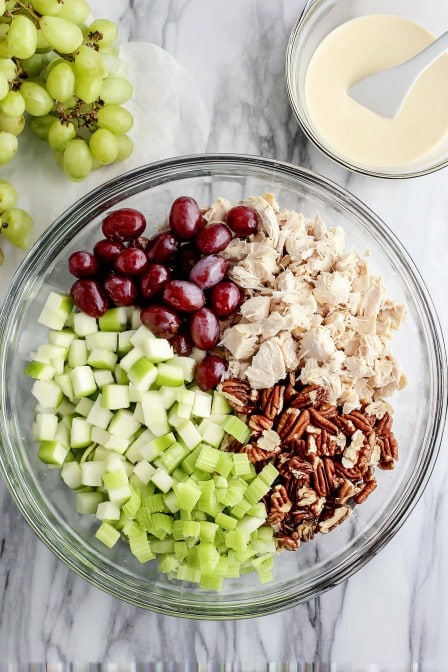 Fresh chicken salad ingredients with grapes, apples, celery, pecans, and dressing in a clear glass bowl.