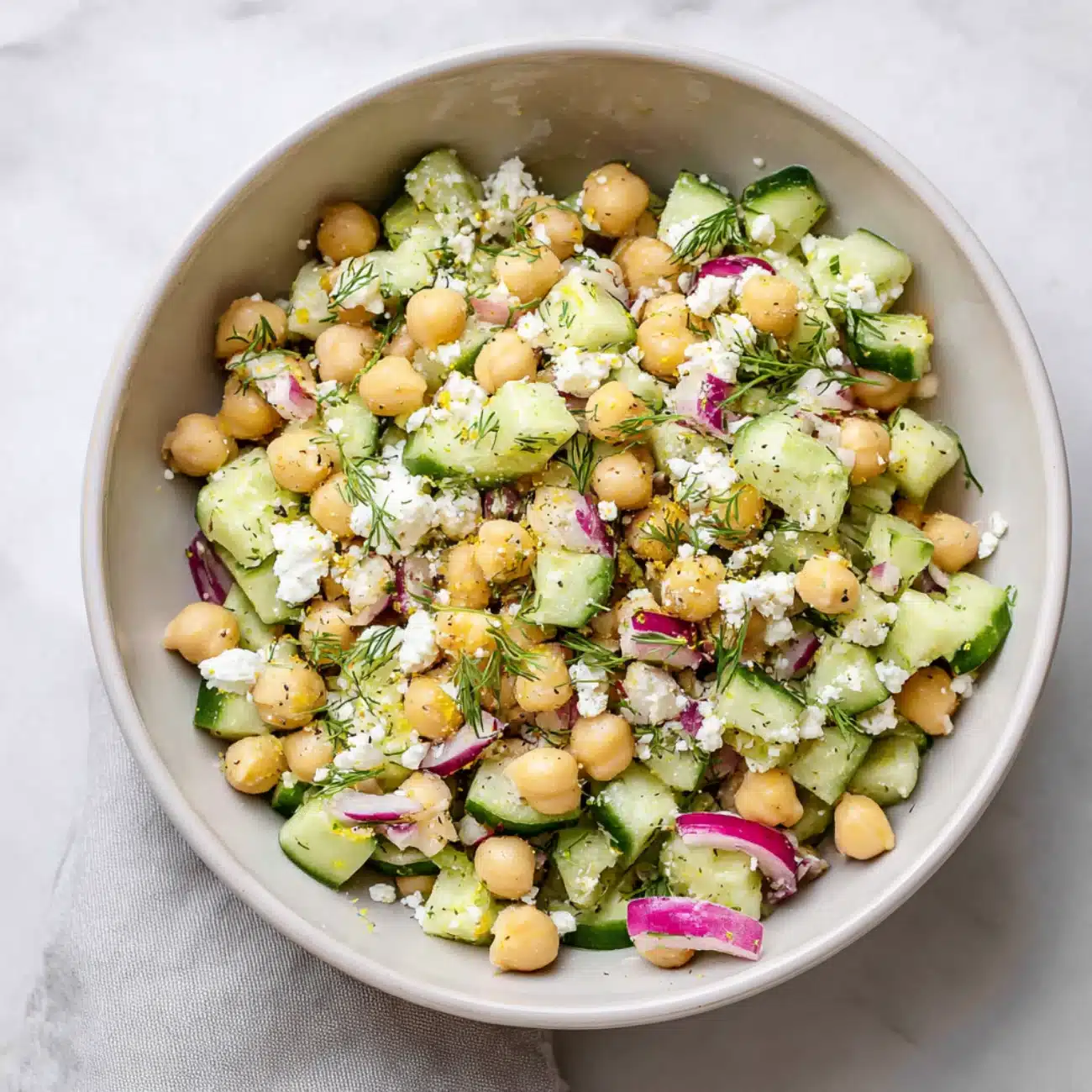 Chickpea cucumber feta salad with dill, red onions, and lemon zest in a beige bowl on a marble surface