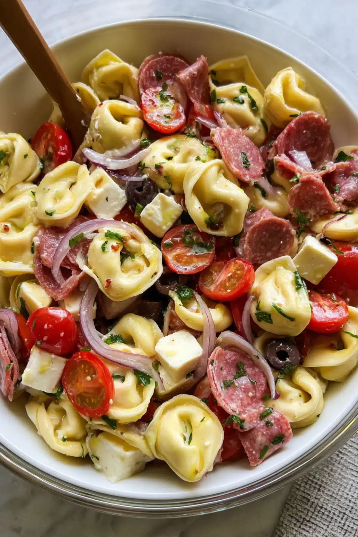 Creamy tortellini Costco pasta salad with salami slices, tomatoes, olives, feta cubes, and red onion in a white bowl.