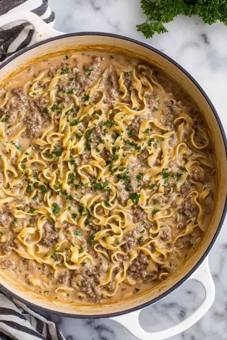 Rich and creamy ground beef stroganoff with egg noodles, garnished with fresh herbs in a white enameled skillet.