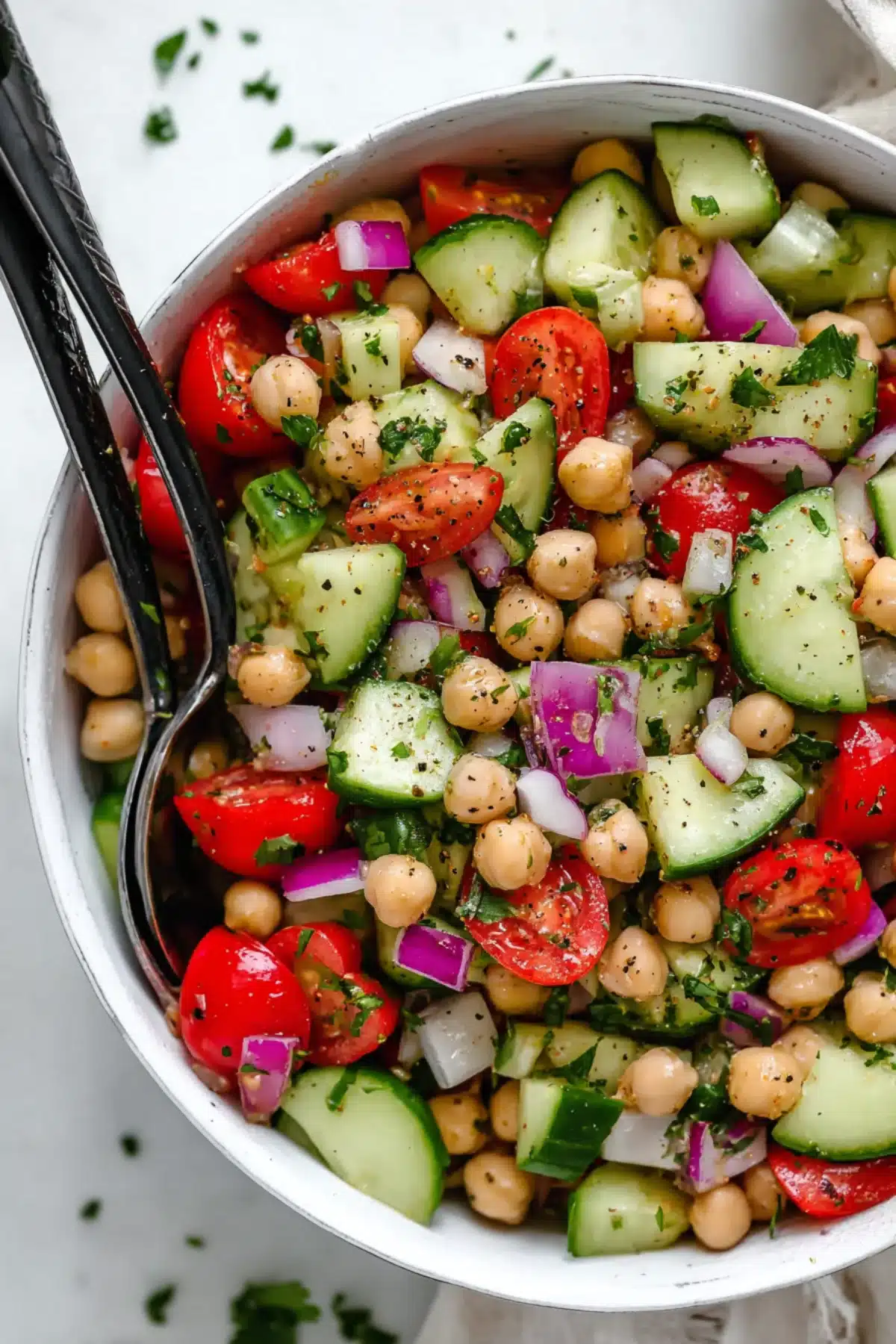 Crisp cucumber, chickpeas, tomatoes, and red onion tossed in herbs and seasoning in a white bowl with black serving spoons