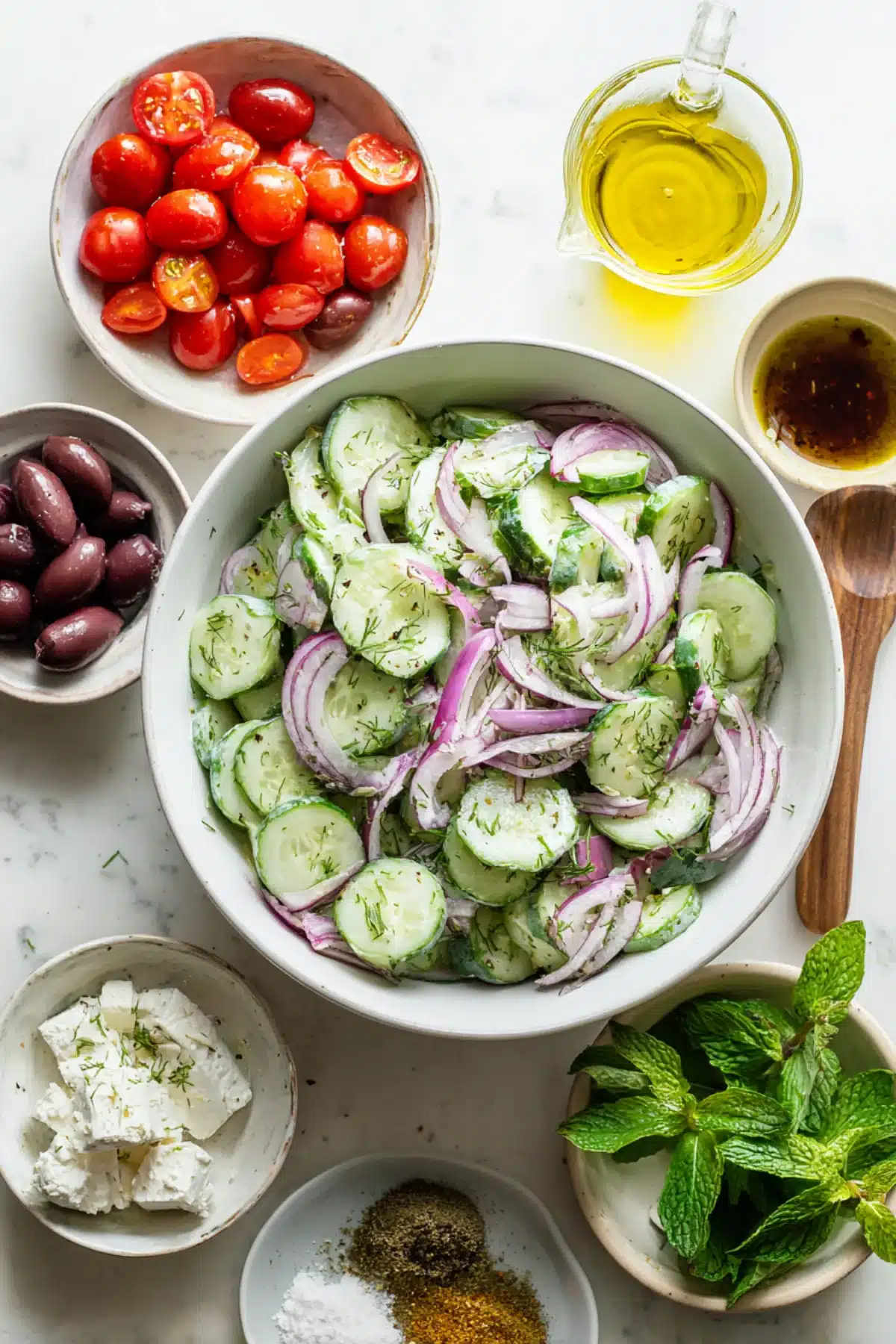 Fresh cucumber salad with red onion, herbs, feta, and Greek yogurt, surrounded by Mediterranean ingredients on white surface