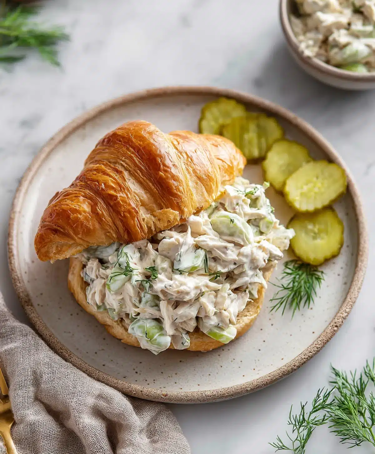 Croissant sandwich filled with creamy dill pickle chicken salad, served with sliced pickles and fresh dill on a plate
