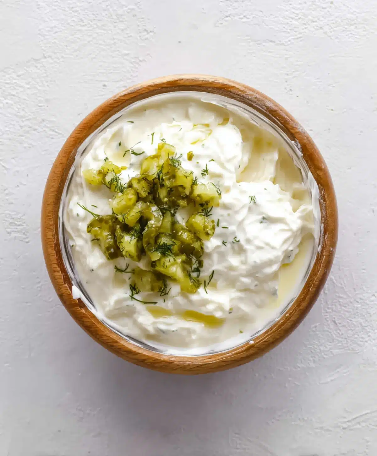 Creamy dill pickle chicken salad dressing with chopped pickles and herbs in a wooden bowl on a white textured surface