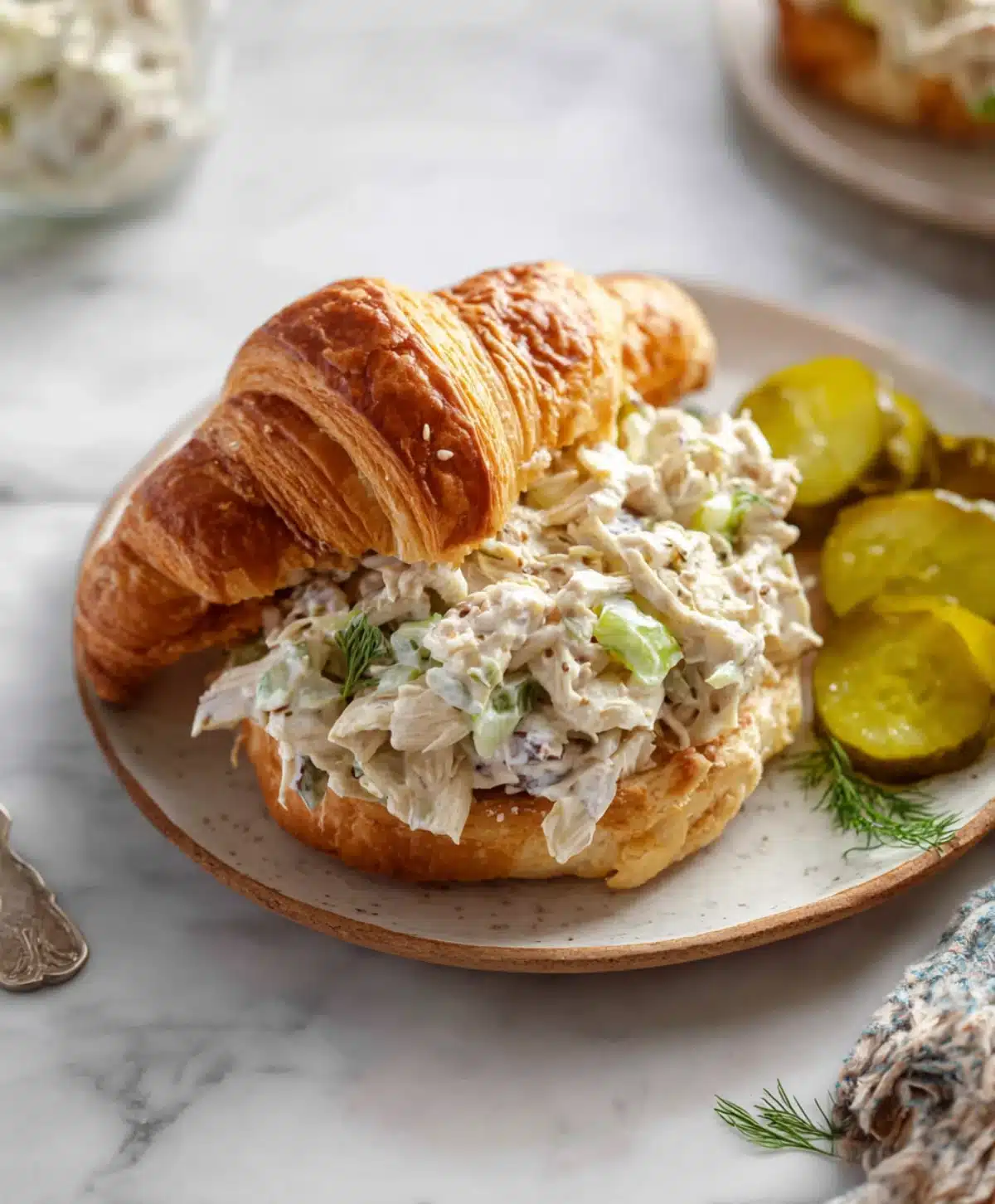 Buttery croissant filled with dill pickle chicken salad served with pickle slices and fresh dill on a ceramic plate