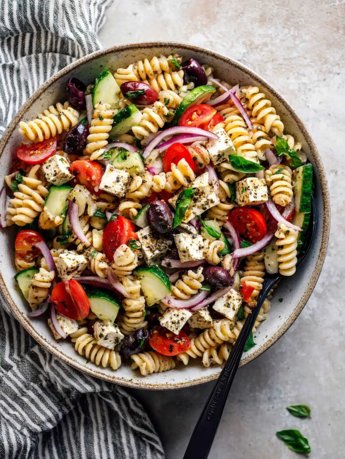 Bowl of high protein pasta salad with feta, cucumbers, tomatoes, red onions, and olives on a neutral stone background.
