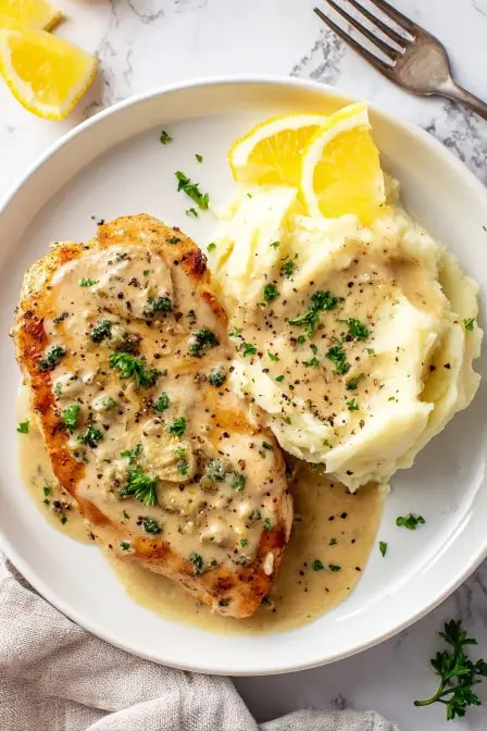 Juicy lemon pepper chicken breast with creamy gravy, served beside mashed potatoes and lemon wedges on a white plate