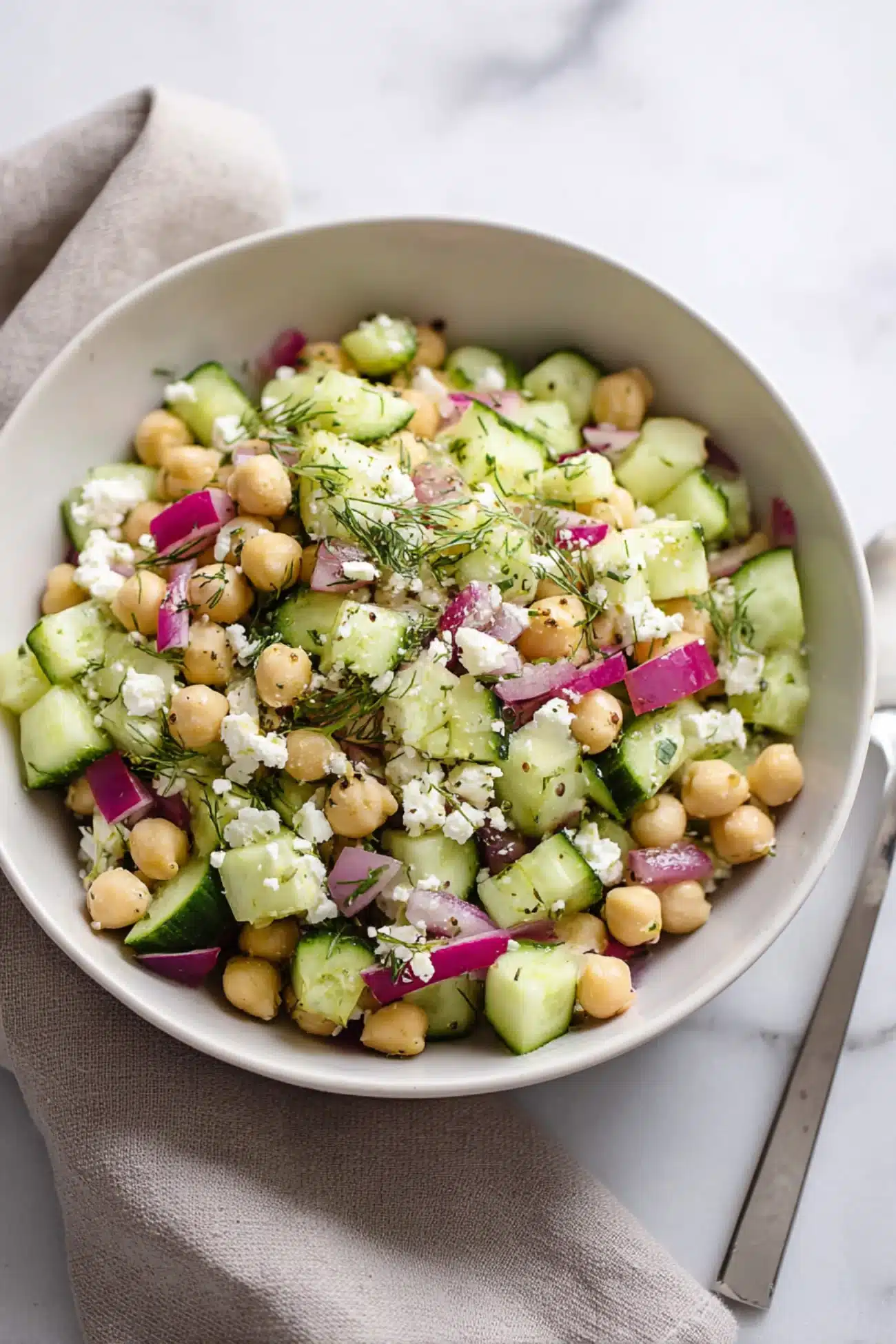 Chickpea cucumber feta salad with fresh herbs, red onions, and olive oil served in a bowl with a napkin and spoon