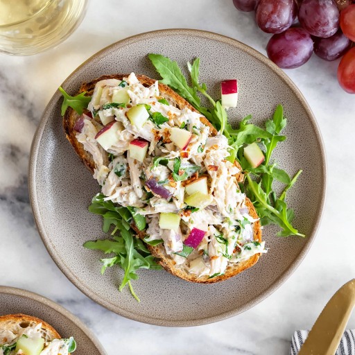 Open-faced chicken salad sandwich with apples, red onion, and arugula on toasted bread, served with grapes on the side
