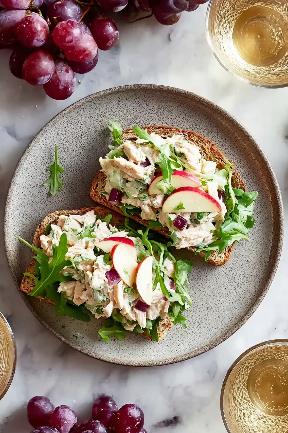 Open-faced chicken salad sandwich with apples, red onion, and arugula on toasted bread, served with grapes on the side