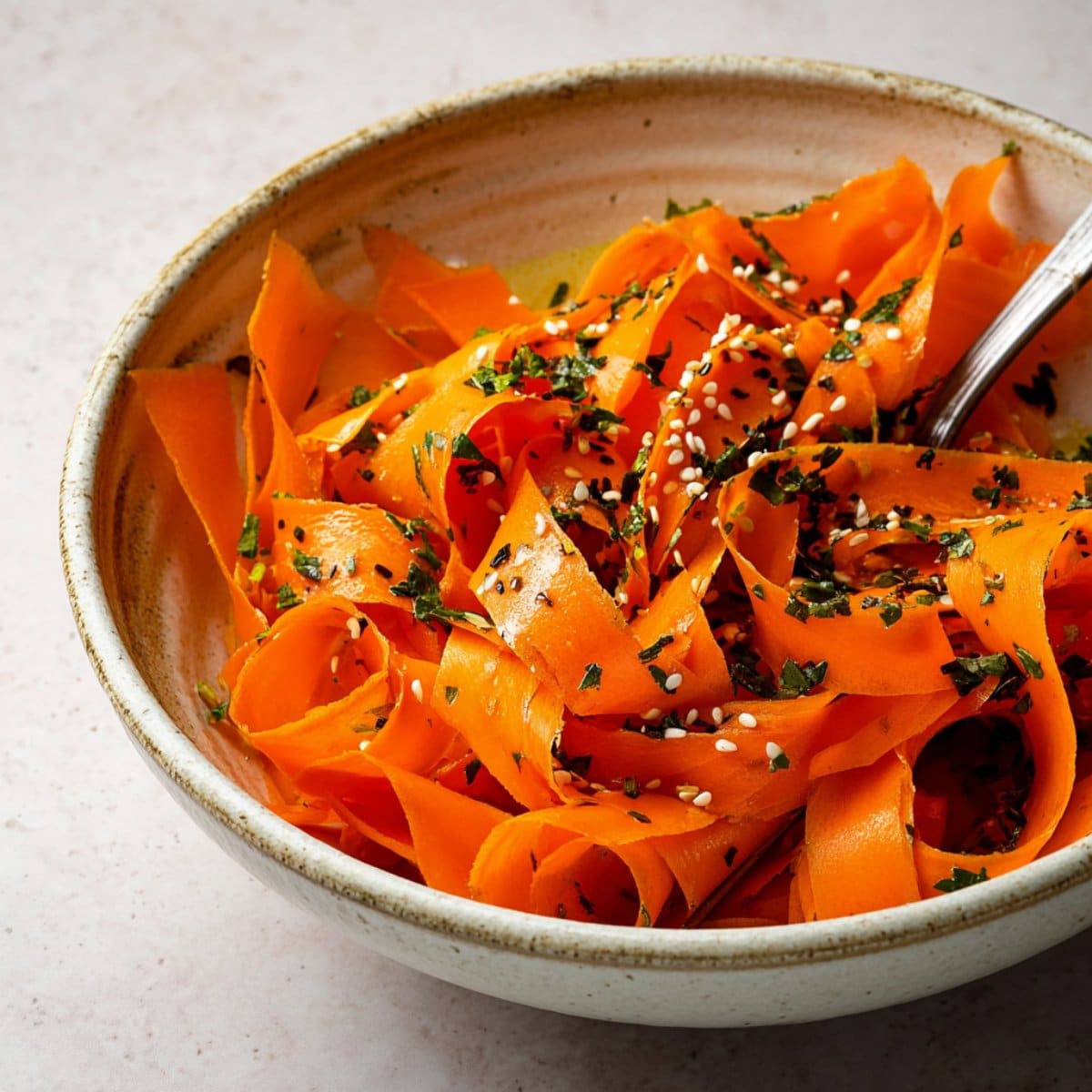 Colorful raw carrot salad with thin ribbons, sesame seeds, and herbs served in a ceramic bowl with a fork.