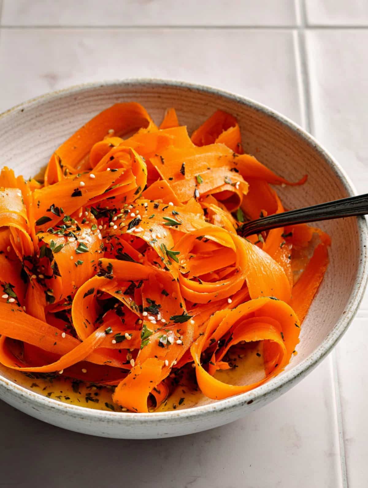 Raw carrot ribbons seasoned with herbs and sesame seeds, beautifully arranged in a bowl on a white tiled surface.