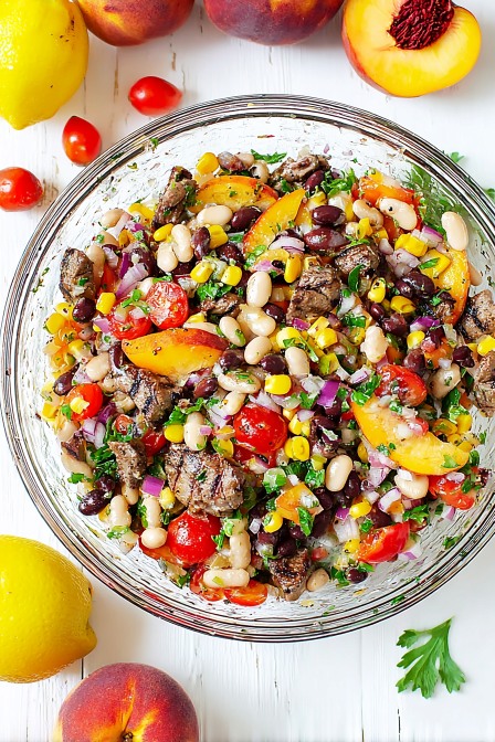 Colorful steak dense bean salad with peaches, corn, cherry tomatoes, red onion, and herbs in a glass bowl
