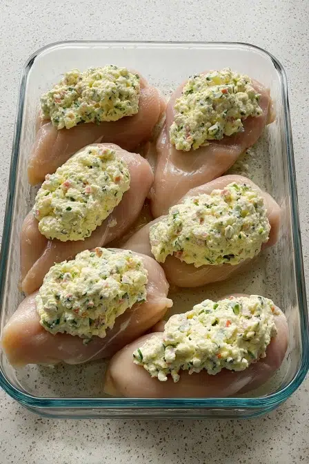 Raw chicken breasts topped with creamy herb and cheese mixture in a glass baking dish, ready for the oven.