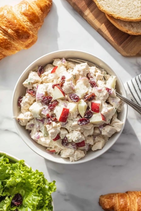 Top view of creamy chicken salad with diced red apples and cranberries in a bowl, surrounded by croissants and bread