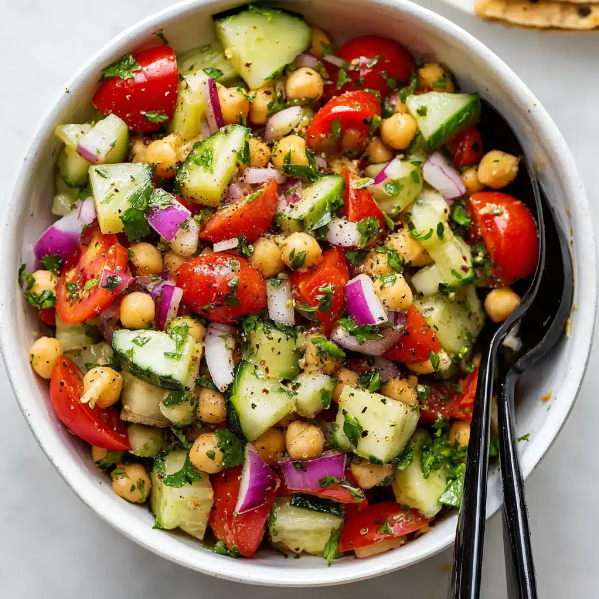 Chickpea cucumber salad with red onion, cherry tomatoes, and herbs served in a white bowl with black spoons