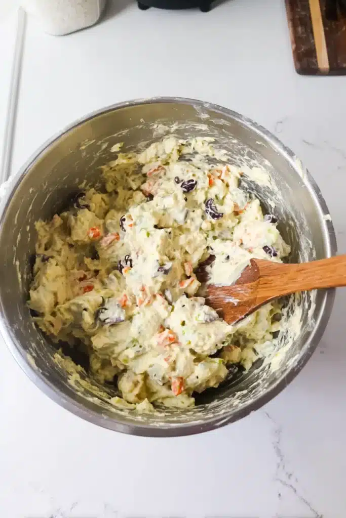 Mixing creamy Amish potato salad with carrots, raisins, and herbs in a stainless steel bowl.