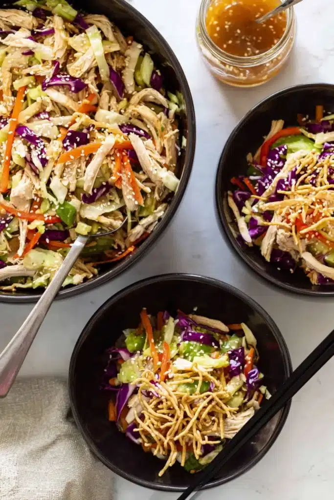 Top view of Chinese chicken salad with shredded chicken, crispy noodles, and sesame seeds in black bowls.