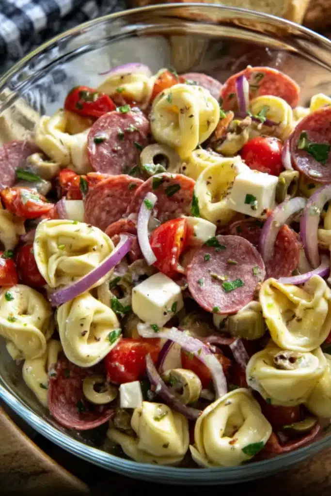 Colorful tortellini pasta salad with salami, cherry tomatoes, olives, mozzarella cubes, and red onion slices in a glass bowl