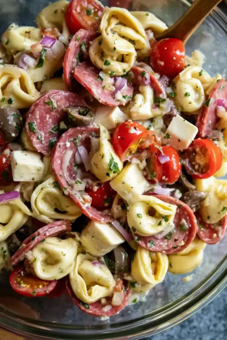Tortellini pasta salad with salami slices, cherry tomatoes, mozzarella cubes, olives, and red onion in a clear bowl