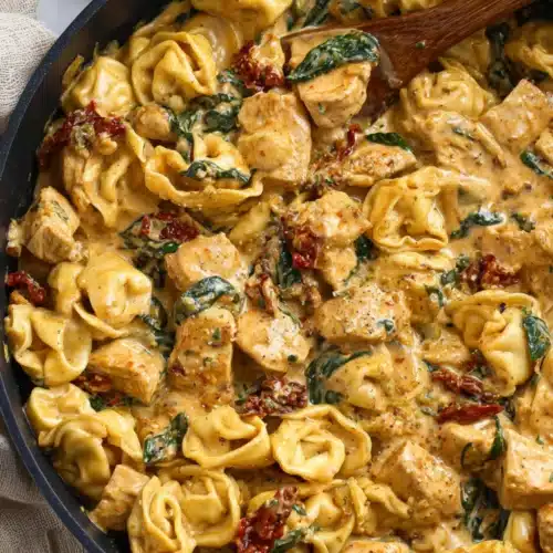 Creamy tortellini pasta with golden chicken cubes, spinach, and sun-dried tomatoes in a rich sauce inside a black skillet