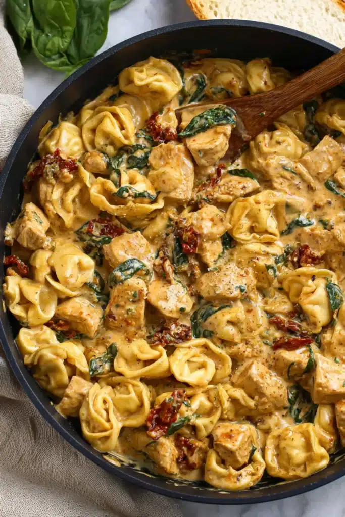 Creamy tortellini pasta with golden chicken cubes, spinach, and sun-dried tomatoes in a rich sauce inside a black skillet