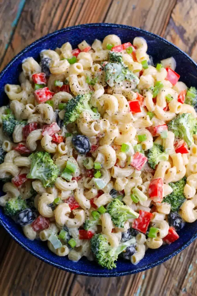  Creamy pasta salad with broccoli, red bell peppers, and black olives in a blue bowl.