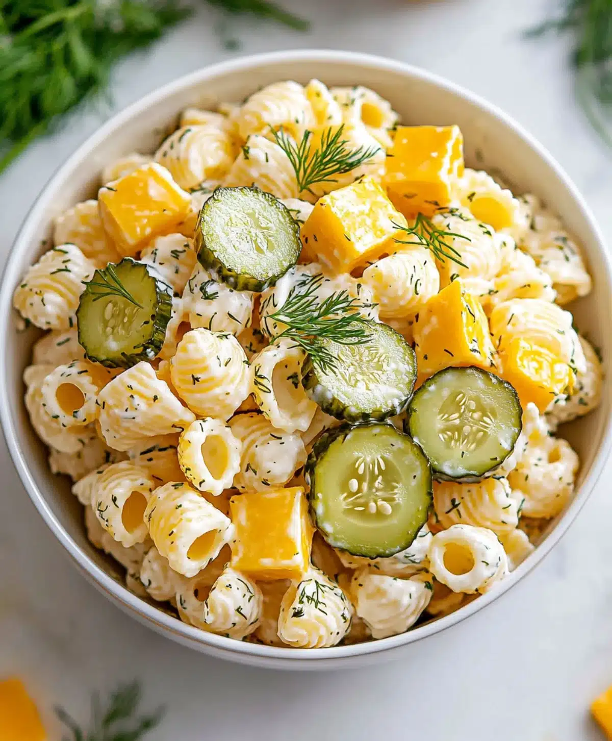 Top-down view of dill pickle pasta salad with shell pasta, cheddar cubes, sliced pickles, and fresh dill garnish