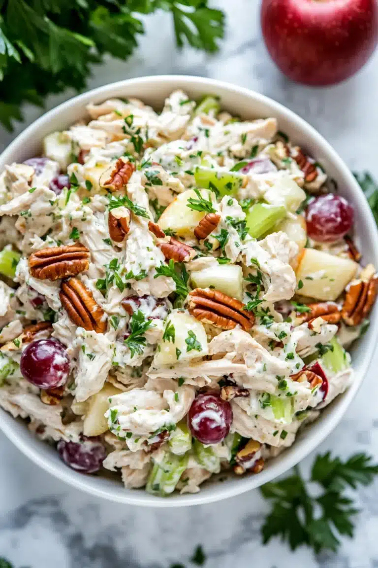 Creamy chicken salad with grapes, apples, celery, toasted pecans, and herbs in a white bowl on a marble surface