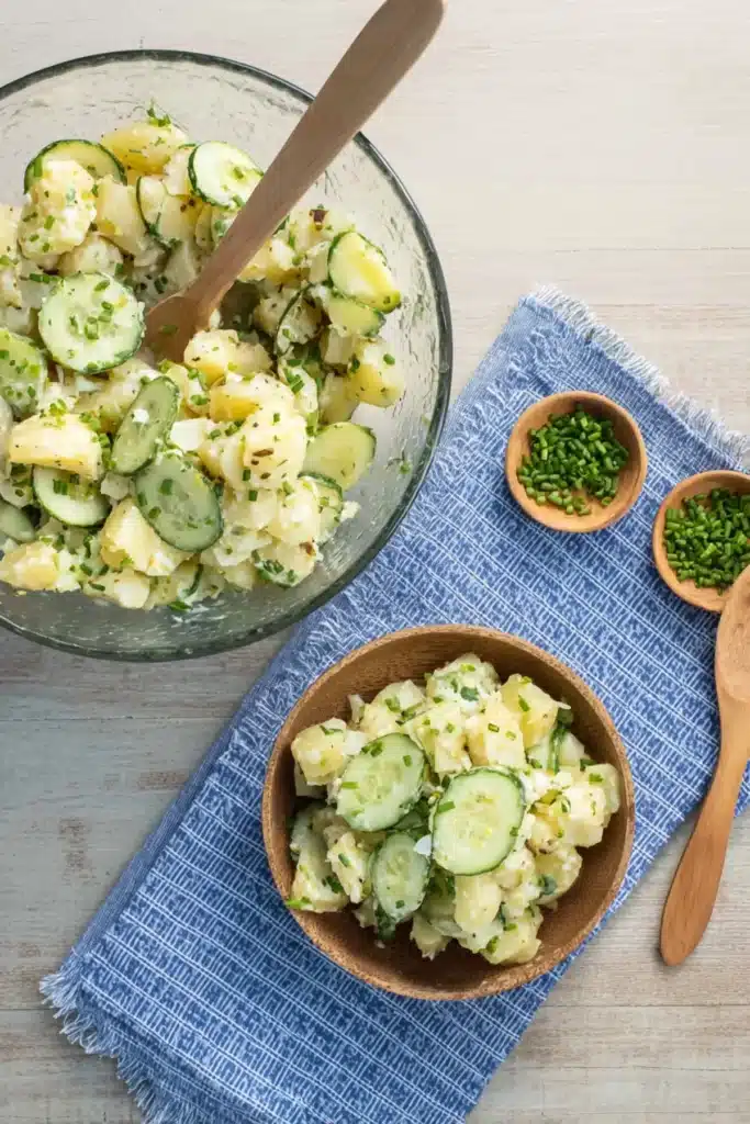 German cucumber potato salad in glass and wooden bowls with sliced cucumbers, potatoes, and chives on a blue napkin