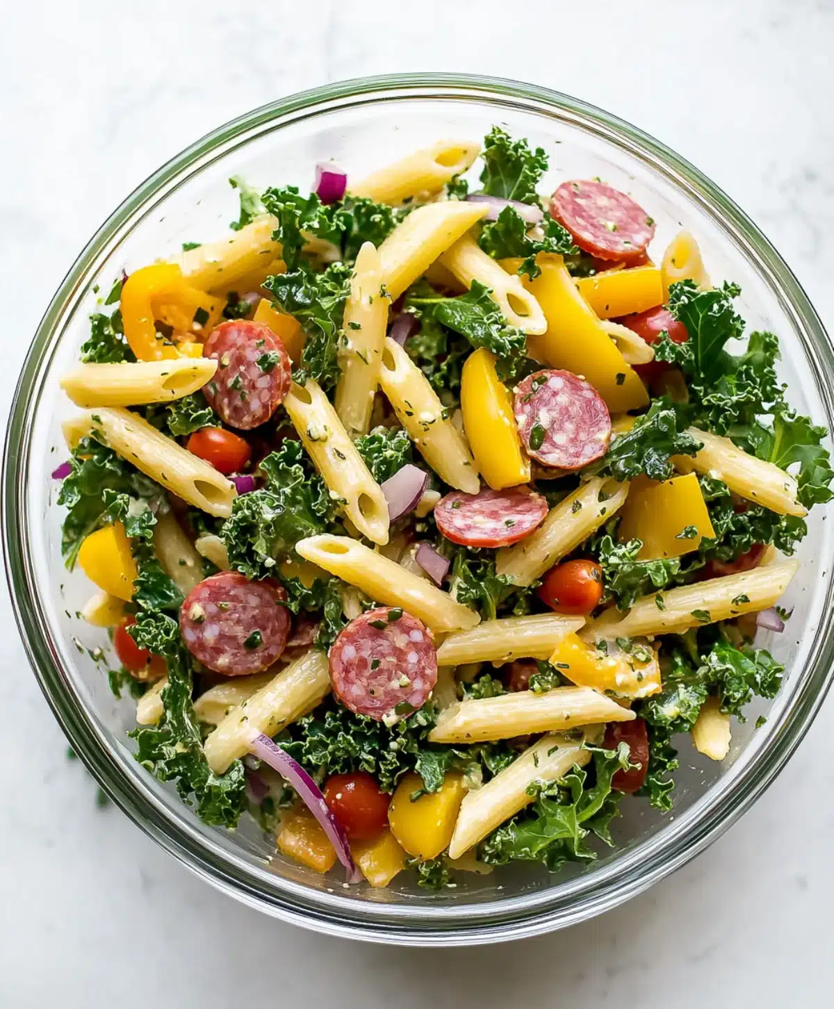 Penne pasta salad with kale, salami slices, yellow bell peppers, cherry tomatoes, and red onions in a glass bowl