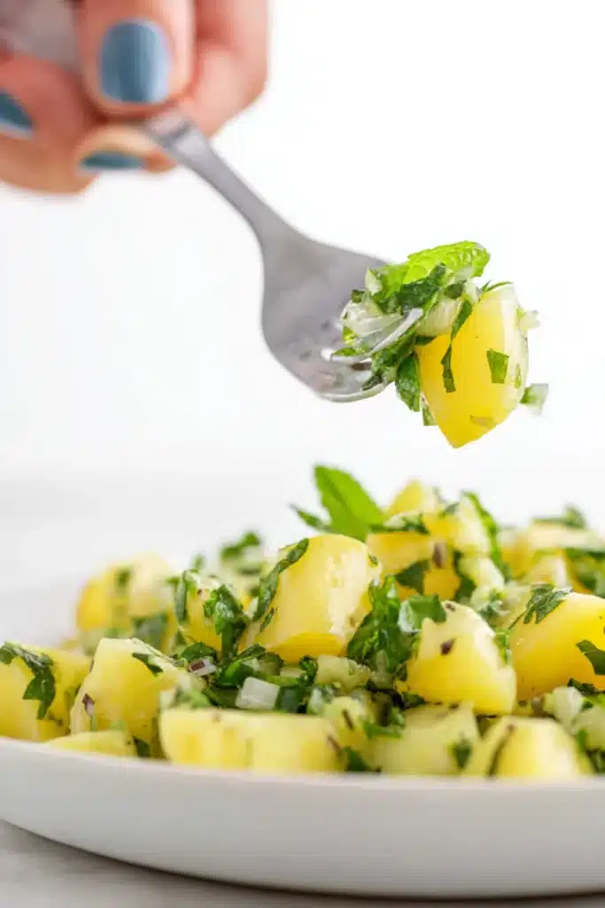 Fork holding bite of Lebanese potato salad with mint, onions, and parsley over a plate of fresh herbed potatoes


