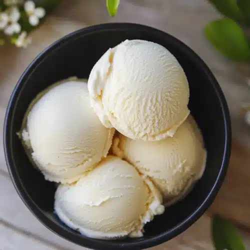 Four scoops of creamy pinon ice cream in a black bowl on a wooden table with white garden flowers around