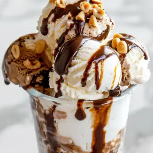 Three scoops of vanilla and chocolate ice cream topped with peanuts and chocolate sauce in a clear glass sundae cup