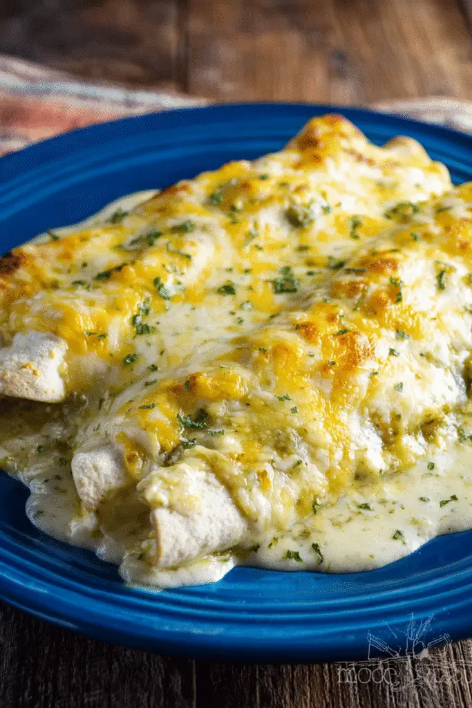 Cheesy sour cream chicken enchiladas topped with golden green chili sauce and melted cheese on a rustic blue plate.