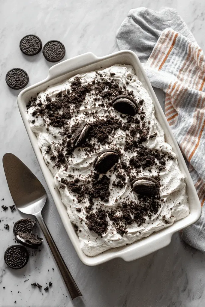Oreo Poke Cake in a white baking dish topped with whipped cream, crushed cookies, and whole Oreos on a marble countertop.