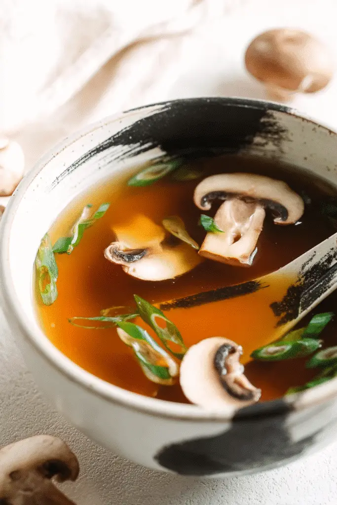 Japanese clear soup with sliced mushrooms and green onions in a black and white ceramic bowl on light background